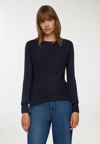 Granatowy sweter dzianinowy z długimi rękami i ściągaczami, z okrągłym dekoltem i luźnym krojem. Noszony z niebieskimi dżinsami.