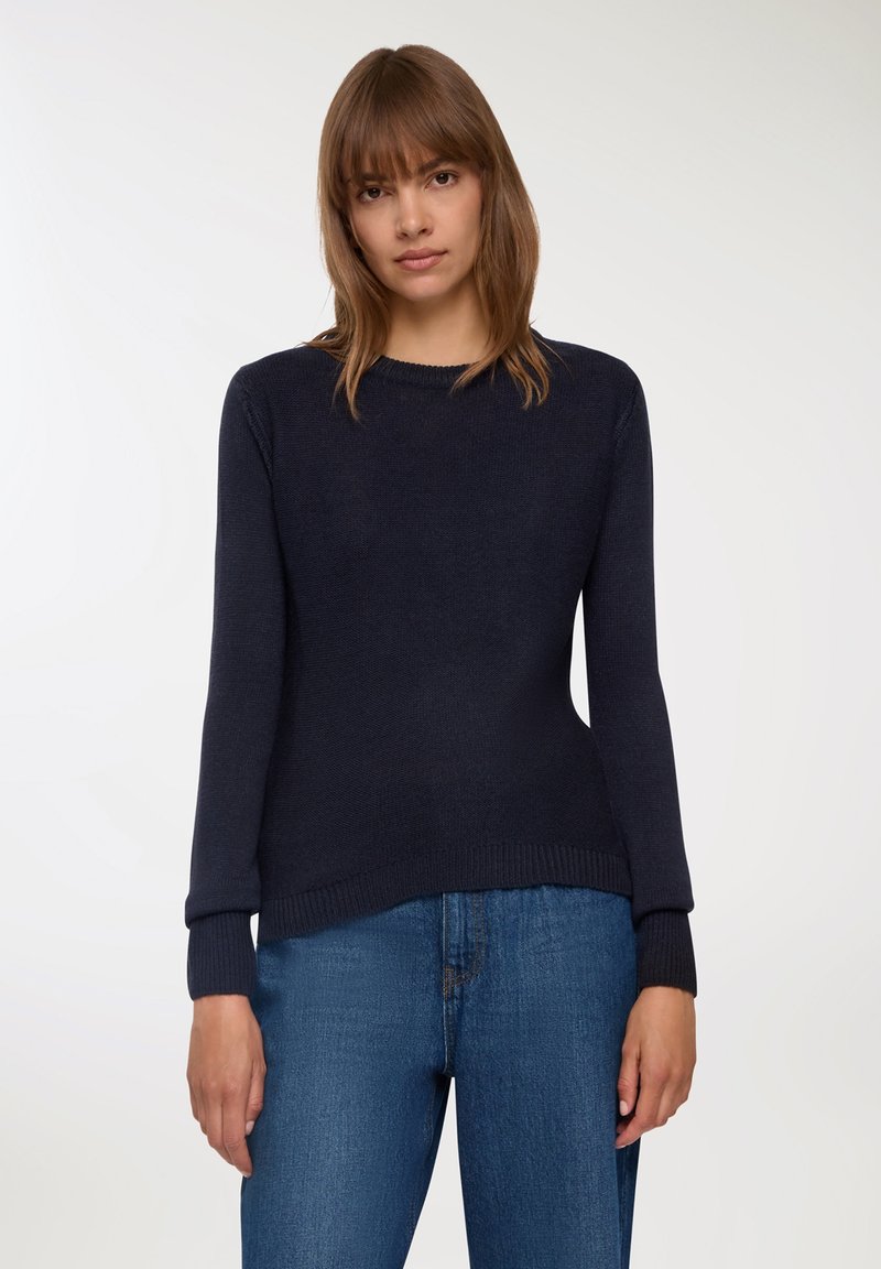 Granatowy sweter dzianinowy z długimi rękami i ściągaczami, z okrągłym dekoltem i luźnym krojem. Noszony z niebieskimi dżinsami.