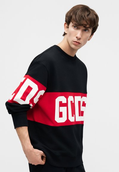 Jaunas vyras su rudos spalvos plaukais, dėvintis juodai raudoną džemperį su didelėmis baltomis raidėmis GCCD ant krūtinės ir rankovės, žiūri į kairę.