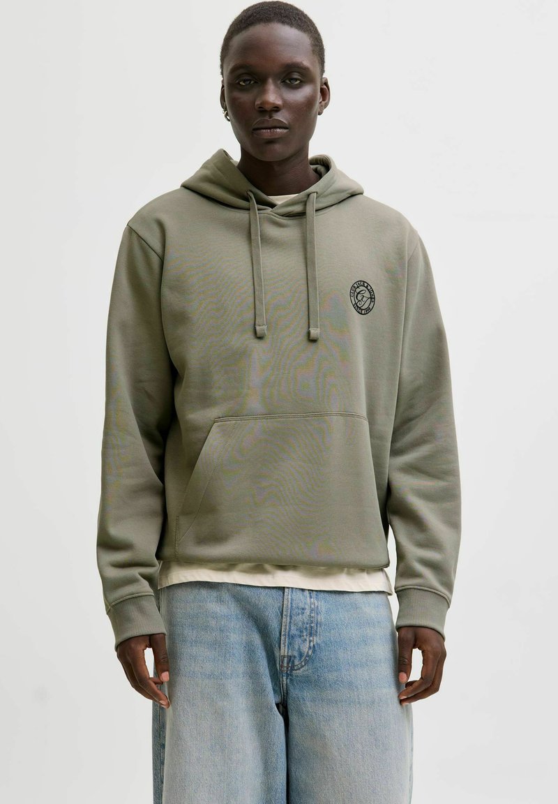 Khaki rohelise kapuutsiga pusa, mis on valmistatud pehmest materjalist, millel on kängurutasku, nöörid ja ümar logomärk rinnal.