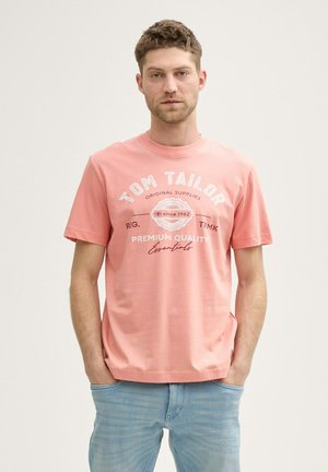 T-shirt en coton rose clair avec des manches courtes et un col rond. Présente un logo et un texte imprimés en blanc sur le devant. Assorti à un jean bleu clair.