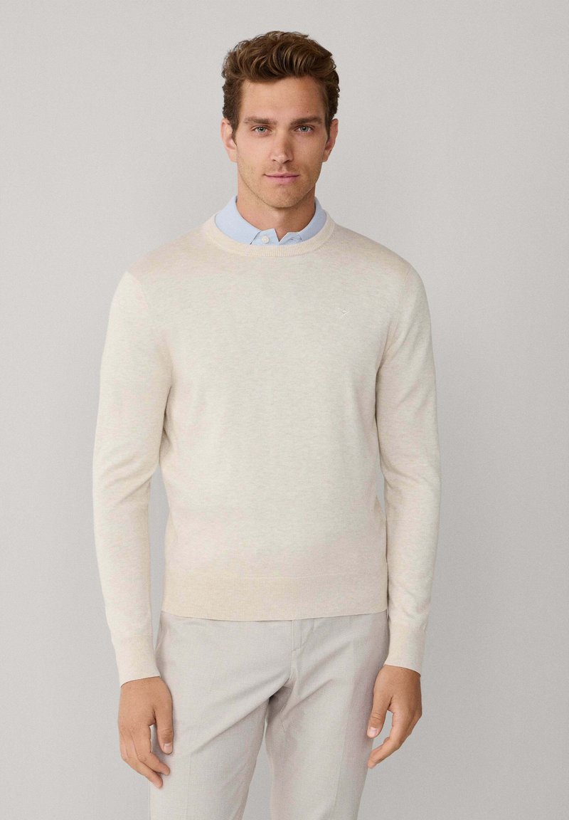Pull en maille beige clair à manches longues, col rond et poignets côtelés, porté par-dessus une chemise bleu clair à col. Texture lisse.