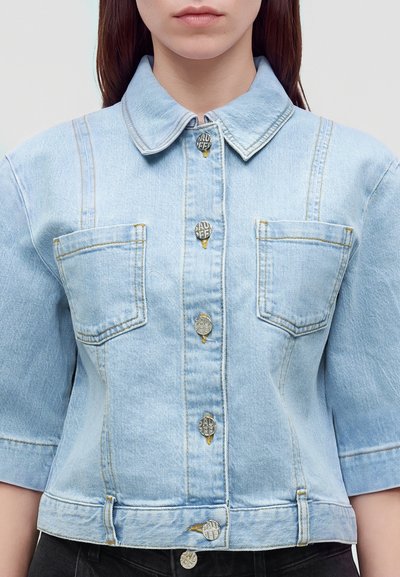 Femme portant une veste en jean bleu clair à manches courtes, deux poches poitrine et des boutons argentés sur le devant.
