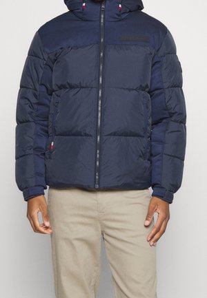 Vue recadrée d’une personne portant une doudoune bleu marine à capuche, zippée sur le devant, et un pantalon beige.