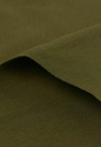 Olivgrüner Stoff mit glatter Textur, der eine leicht gefaltete Kante aufweist, die ein feines geripptes Muster zeigt. Weiche und matte Oberfläche.