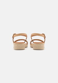 Sandales en cuir couleur tan avec des brides tressées, une semelle compensée basse et une semelle intérieure texturée et mouchetée. Fermeture à boucle réglable au niveau de la cheville.