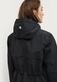 Veste imperméable noire avec capuche, taille ajustable par cordon, et un petit logo brodé dans le dos. Tissu lisse et léger.