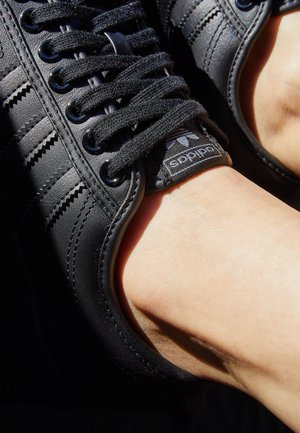 Zbliżenie na czarny sneaker marki Adidas z grubymi sznurówkami, noszony na stopie w jasnym świetle słonecznym, ukazujący teksturowaną skórę i detale logo.