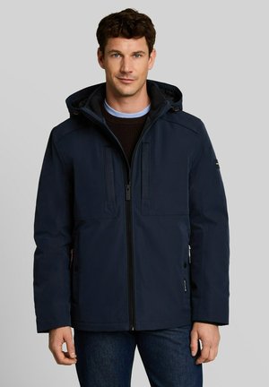 Man draagt een marineblauw gewatteerd jack met capuchon, dichtgeritst over een donkere trui en lichtblauw overhemd, staand tegen een effen grijze achtergrond.