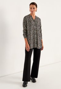 Bluse mit Blumenmuster, V-Ausschnitt, langen Ärmeln und Knopfdetails. Kombiniert mit schwarzen Schlaghosen und robusten schwarzen Schuhen.