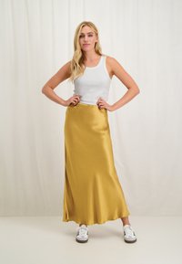 Gele satijnen maxi rok met een gladde textuur, gecombineerd met een wit geribbeld tanktop en witte sneakers. Model poseert met de handen op de heupen.