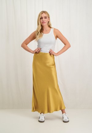 Gele satijnen maxi rok met een gladde textuur, gecombineerd met een wit geribbeld tanktop en witte sneakers. Model poseert met de handen op de heupen.
