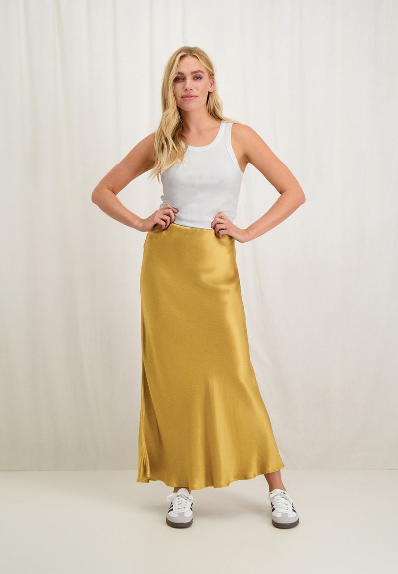 Gele satijnen maxi rok met een gladde textuur, gecombineerd met een wit geribbeld tanktop en witte sneakers. Model poseert met de handen op de heupen.