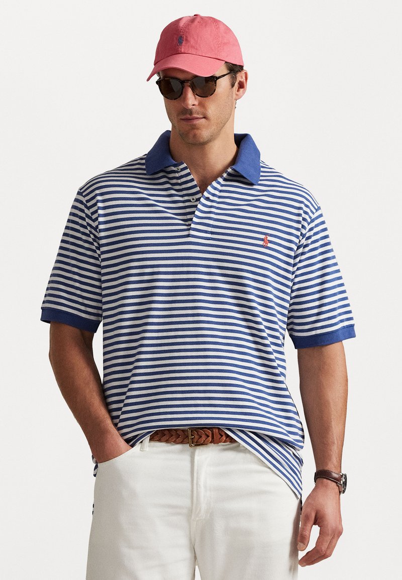 Vīrietis brunā un baltā svītrotā polo krekla ar zilu apkakli, baltās biksēs, brūnu pītā jostā, rozā cepurē, saulesbrillēs un rokas pulkstenī.