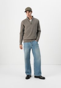 Bruin gebreid sweater met een halve rits, lichtblauwe wijde jeans en een hoed met ruitpatroon. Zwarte enkellaarsjes maken de outfit compleet.