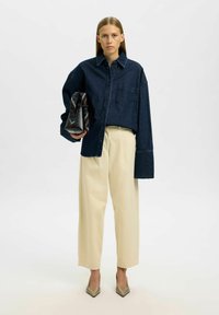 Dunkelblaue weite Denim-Bluse mit einer Brusttasche, cremefarbene Hose mit weitem Bein und spitze beigen Absätze, während sie eine schwarze strukturierte Tasche hält.