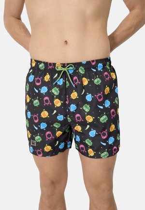 Bañador corto negro para hombre con estampados de monstruos de dibujos animados coloridos y cintura con cordón verde, usado por una persona sin camiseta.