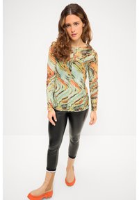 Langärmliges Oberteil mit einem marmorierten Muster in Grün-, Orange- und Beigetönen, kombiniert mit schwarzen Leggings und beigefarben-orangefarbenen Stiefeln.