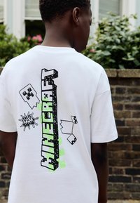 Un garçon vu de dos portant un t-shirt blanc de Minecraft avec des visages de Creeper pixelisés et des graphiques de blocs TNT en extérieur.