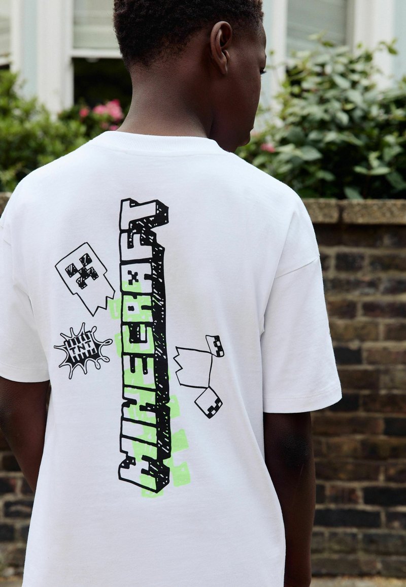 Un garçon vu de dos portant un t-shirt blanc de Minecraft avec des visages de Creeper pixelisés et des graphiques de blocs TNT en extérieur.
