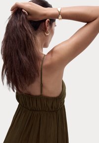 Olivegrünes, plissiertes Kleid mit dünnen Trägern, geraffter Taille und offenem Rücken. Dazu goldene Creolen und ein gehämmertes Goldarmband.