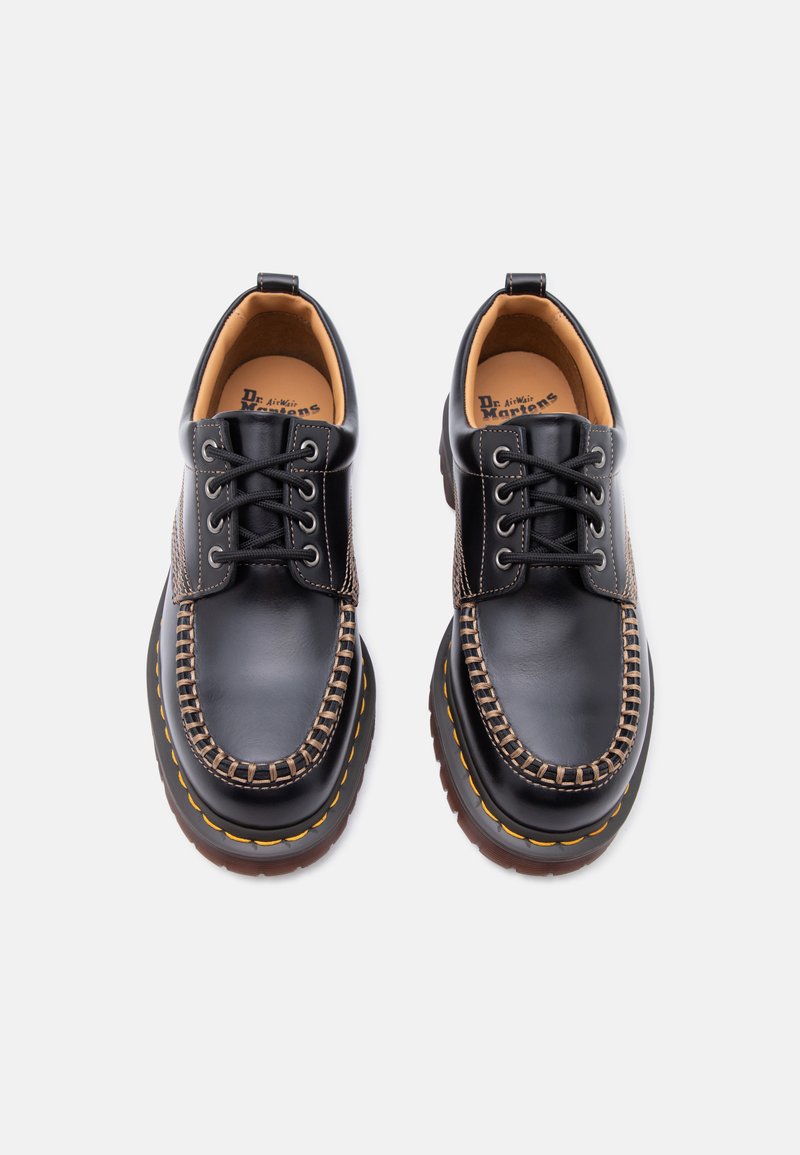 Schwarze Dr. Martens Lederschuhe mit dicken, gelb abgesteppten Sohlen und Mokassin-ähnlicher Schnürung, von oben auf weißem Hintergrund dargestellt.