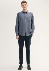 Camisa azul de manga larga con cuello, bolsillo en el pecho, y cierre de botones al frente, combinada con pantalones oscuros y zapatos negros.