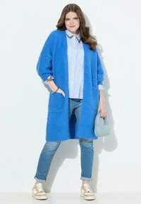 Frau, die einen blauen, flauschigen langen Cardigan, ein gestreiftes Shirt, hochgekrempelte Jeans und beige Sneakers trägt, während sie eine kleine hellblau Handtasche hält und vor einer weißen Wand steht.