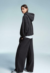 Ung kvinde iført en oversized sort hættejakke, brede bukser, hvide sneakers og en hvid skjorte, stående med hænderne i lommerne og kigger over skulderen.