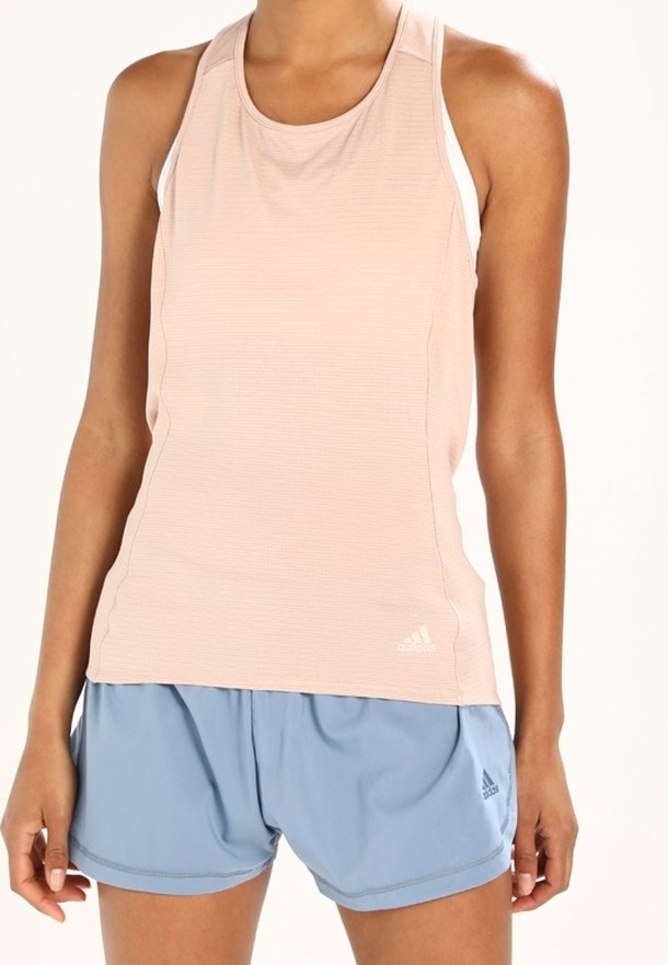 Femme portant un débardeur sportif rose pâle et un short Adidas bleu clair, debout devant un fond uni.