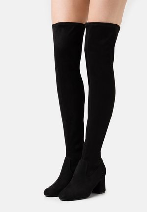 Černé semišové kozačky nad kolena s blokovými podpatky, nošené na bosých nohách, zobrazené zepředu na jednobílém pozadí.