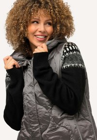 Chaleco acolchado en gris con un acabado acolchado, que presenta mangas de punto negras adornadas con un acento de patrón blanco y gris. Cierre frontal con cremallera.