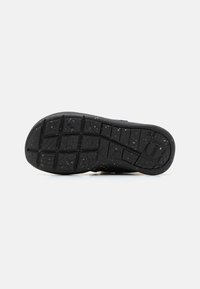 Suela de zapatilla negra con un patrón de relieve texturizado y pequeñas motas blancas. Presenta un borde curvado y una sección elevada con el logo de la marca.