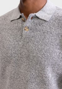 Grå teksturert polo shirt med et mykt strikket stoff, med kragehals, to knapper foran og synlige sømmer.
