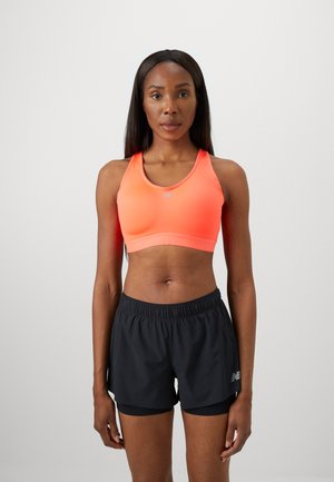 Femme athlétique se tient de face, portant un soutien-gorge de sport corail et un short de course noir en couches, sur un fond uni.