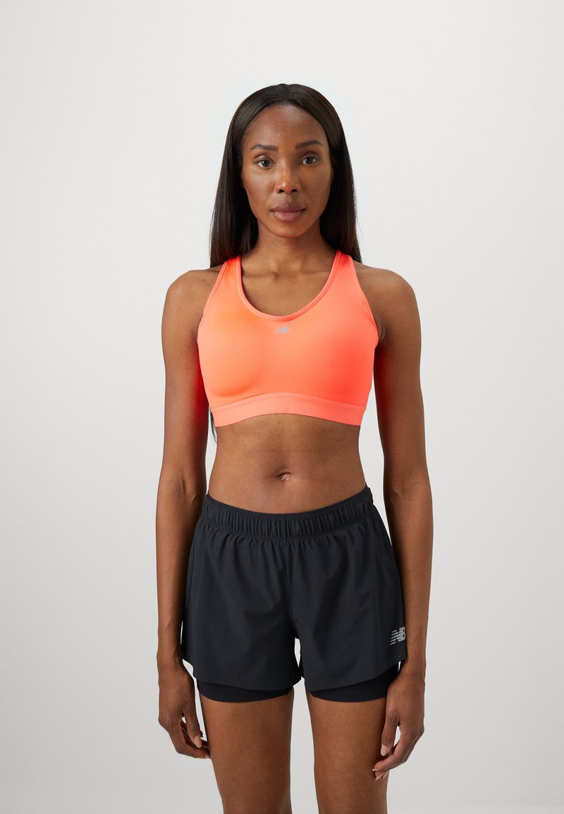Una mujer atlética está de pie de frente, vistiendo un sujetador deportivo color coral y pantalones cortos de running negros en capas, frente a un fondo liso.