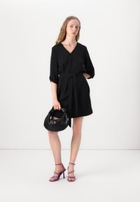 Schwarz kurzes Kleid mit V-Ausschnitt und Gürtel, Dreiviertelärmel. Das Model hält eine schwarze Handtasche und trägt lila Riemchen-Heels.