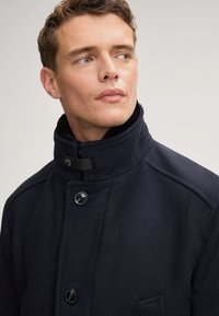 Cappotto in misto lana blu navy con colletto alto, dotato di due bottoni, chiusura con zip e una finitura testurizzata. Dettagli sottili sulle spalle.