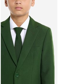 Giacca verde con tessuto strutturato e revers a punta, con chiusura a un bottone, indossata sopra una camicia bianca e una cravatta abbinata.