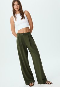 Pantaloni larghi verde oliva con pieghe, dotati di passanti per cintura e dettaglio fibbia. Abbinati a un fitted crop top bianco.