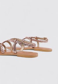 Sandalias planas con un diseño de tira trenzada metálica en rosa, hebilla ajustable y una planta de cuero marrón suave. Estilo simple y minimalista.