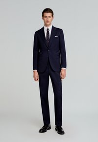 Costum pe navy, realizat la comandă, cu un sacou cu un singur rând de nasturi și două buzunare, purtat cu o cămașă albă și o cravată cu model. Pantofi negri, lustruiți.