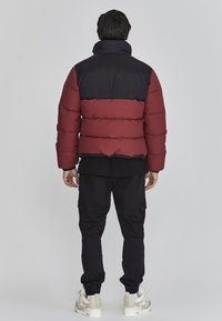 Chaqueta de plumas en rojo y negro, con secciones acolchadas horizontales, cuello alto, llevada sobre una camiseta negra, combinada con pantalones cargo negros y zapatillas.