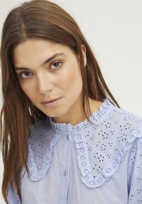 Svetlo modra bluzica z vezenimi detajli, nagubanim ovratnikom, sprednjim gumbom in nežnimi odprtimi vzorci na ramenskem in rokavih.