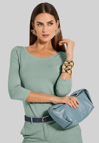 Mintgroene lang mouwtop, marineblauwe textuurriem, gouden chunky armband en blauwe leren clutch. Duidelijke oversized oorbellen en glad haar.