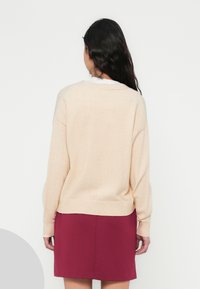 Ανοιχτό μπεζ πουλόβερ με άνετη εφαρμογή, ριμπ μανσέτες και τελείωμα, σε συνδυασμό με σκούρα μπορντό φούστα. Μαλακή υφή, μινιμαλιστικός σχεδιασμός, χωρίς ορατά σχέδια.