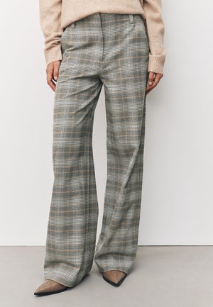 Mujer con pantalones de cuadros de talle alto y pierna ancha en tonos grises y beige, combinados con un suéter beige y zapatos marrones de punta puntiaguda.