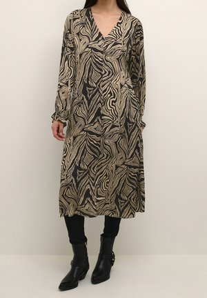 Kvinde iført en langærmet, V-hals kjole med abstrakte sorte og beige swirl mønstre, kombineret med sorte leggings og ankelstøvler.