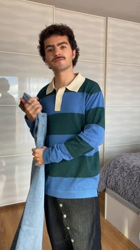 Jeune homme avec moustache tenant un jean plié, portant un t-shirt à manches longues rayé bleu et vert, debout dans une chambre avec un sol en bois.