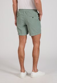 Hombre con pantalones cortos verde claro con cintura elástica y bolsillos traseros con botones, zapatillas blancas, de pie sobre fondo liso.